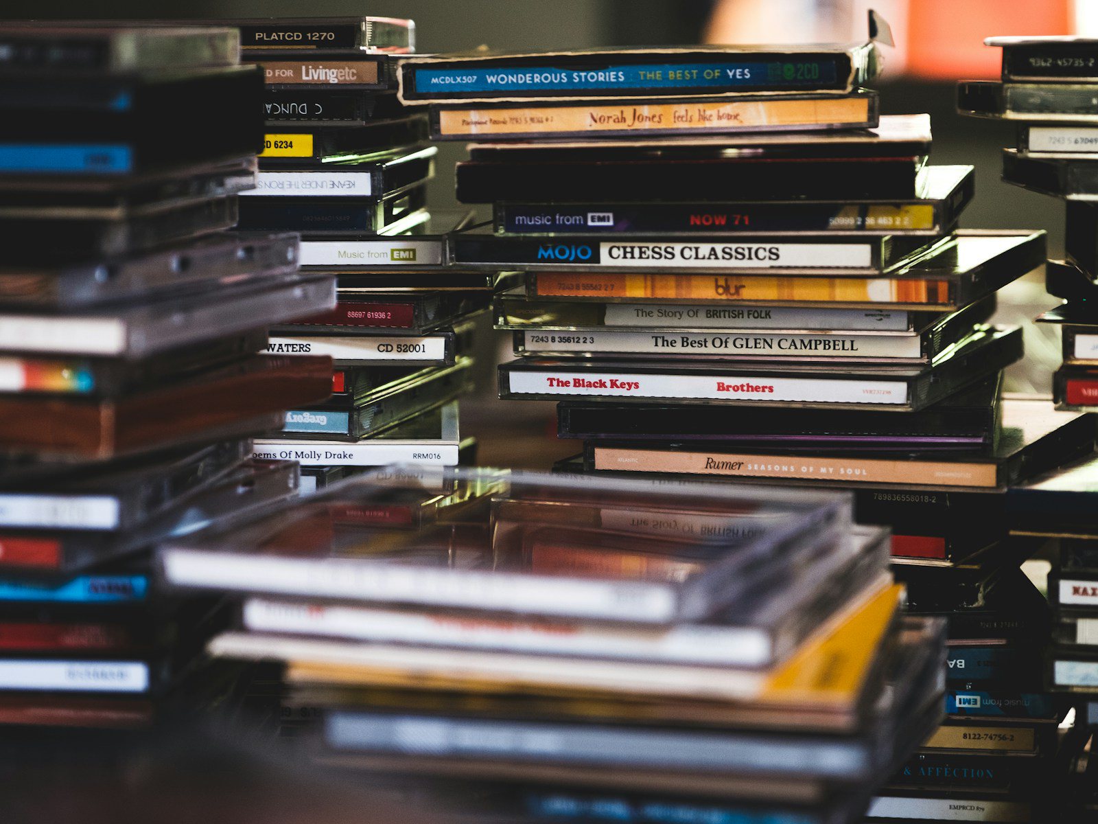Warm desk scene with a short stack of CD cases, a phone face up showing abstract album art, and a pair of earbuds, no people