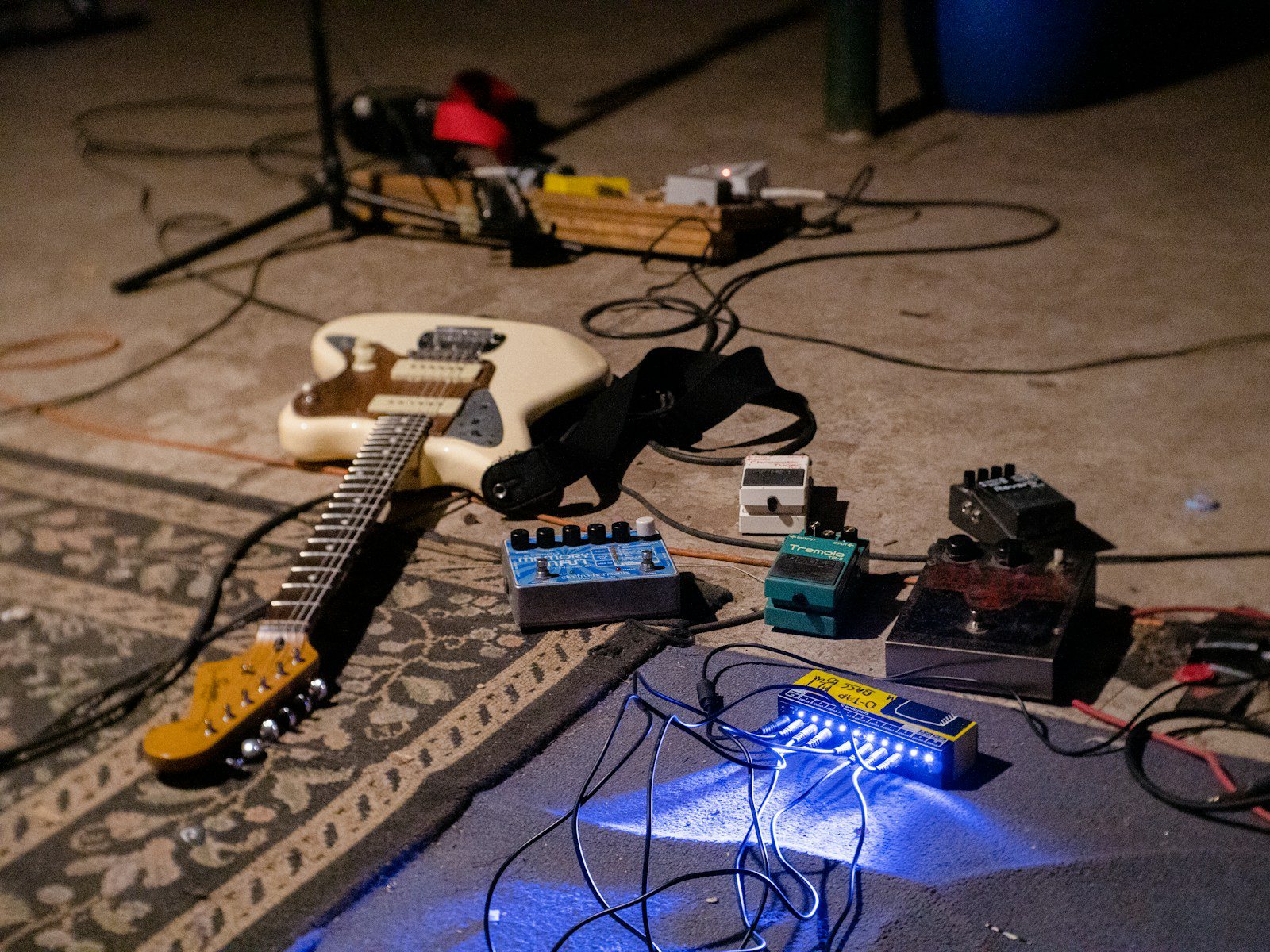Effects pedal with a footswitch and three knobs lit by a single beam of colored light on a dark stage floor, no people
